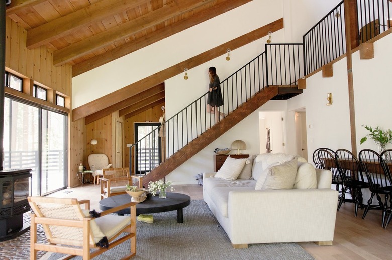 A woman descending a wood staircase. A living room with eclectic furniture, stove fireplace, stone hearth, and wood beam ceiling.