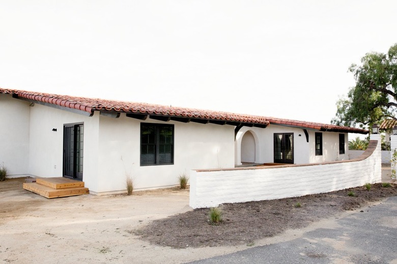 Main and side entrances of a white Spanish-style home with a clay tiled roof. A white brick perimeter wall runs in front of the home. An archway leading to the front door is visible. Two wooden steps lead up to the side entrance.