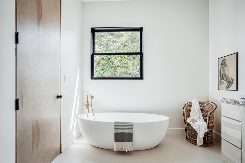 Minimalist bathroom with light wood door, white walls and floors, freestanding bathtub with towel, peacock chair with white towel