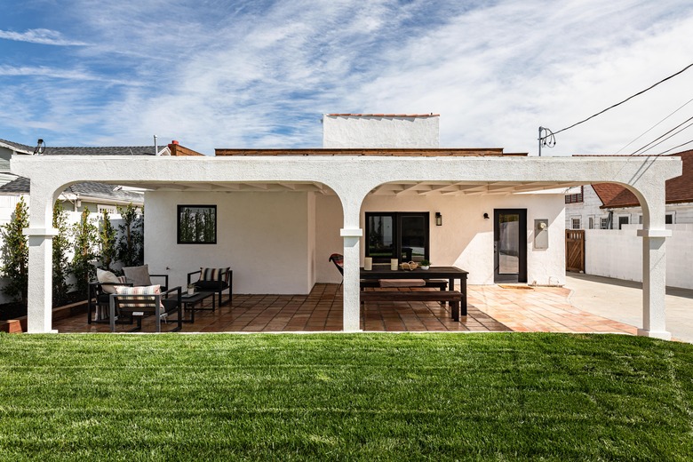 The backyard of a Spanish-style home; green grass in the yard and patio furniture underneath the covered patio