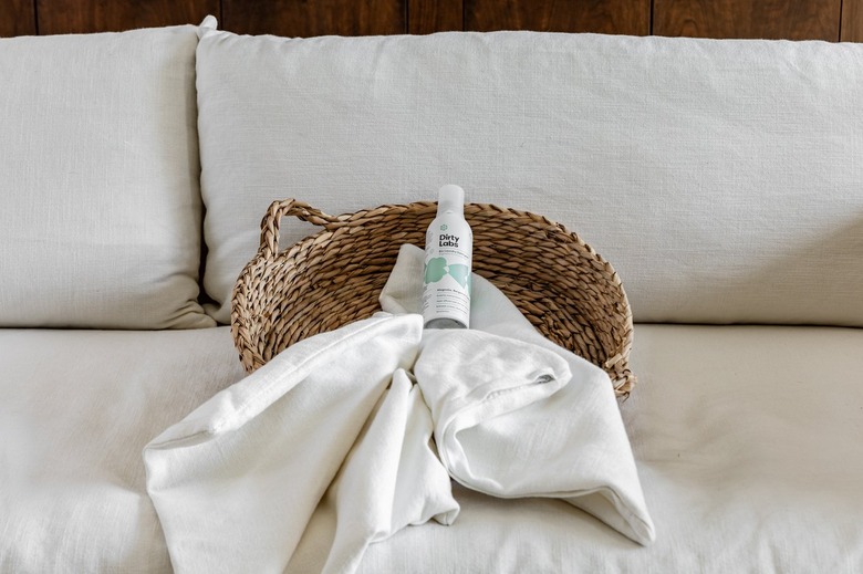 A laundry basket on a white couch with laundry detergent and a cushion cover in it.