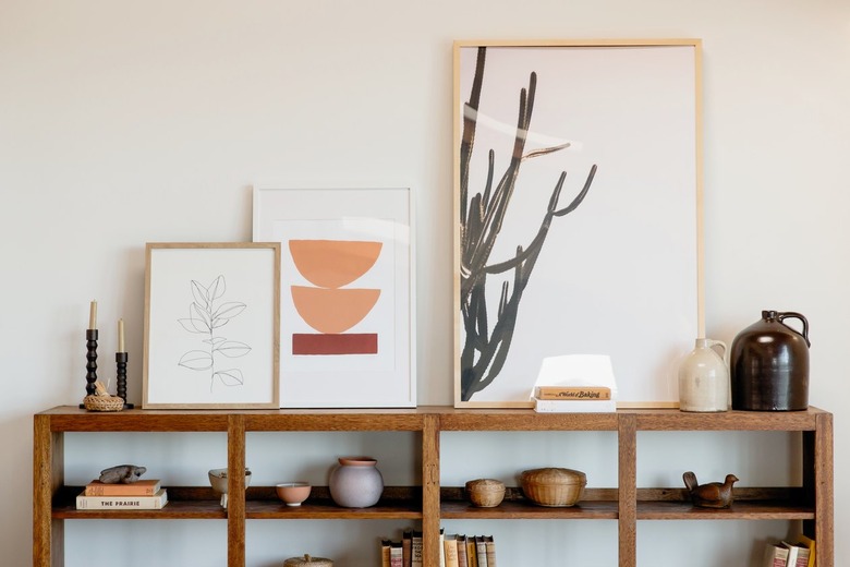 Wooden bookcase against a white wall. On its shelves, books and other decorative trinkets such as ceramic jugs, woven baskets, and a wooden dove figurine. On top, two candlestick holders and tree pieces of framed art.