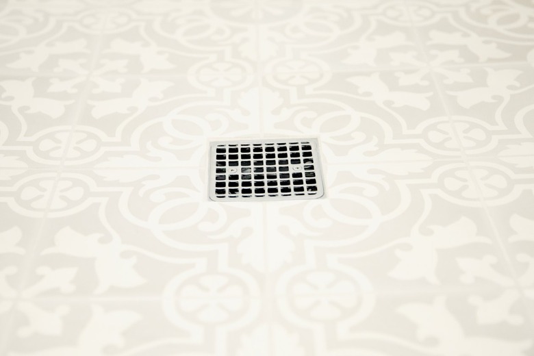 Closeup of white and grey patterned bathroom floor tiles with a chrome square grille shower drain in the middle.