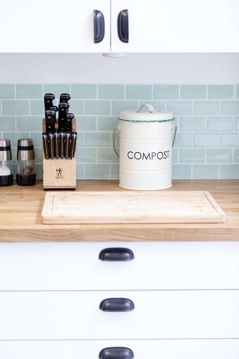 Kitchen counter with knife set, salt and pepper shakers, and compost bucket on wood countertop and white shelves against tiled backsplash