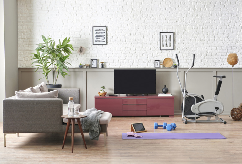 Man is doing sport and looking cell phone training program. Decorative living room, television unit, grey sofa purple mat and blue dumbbell brick wall home interior concept.