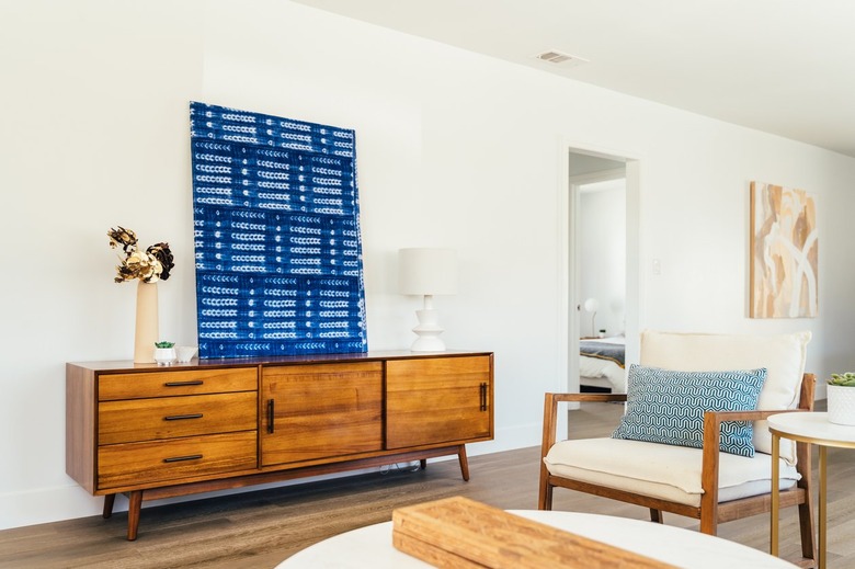 Wood sideboard with a contemporary art piece, dry flowers in vase, a white lamp. Wood armchair with white cushions and blue pillow.