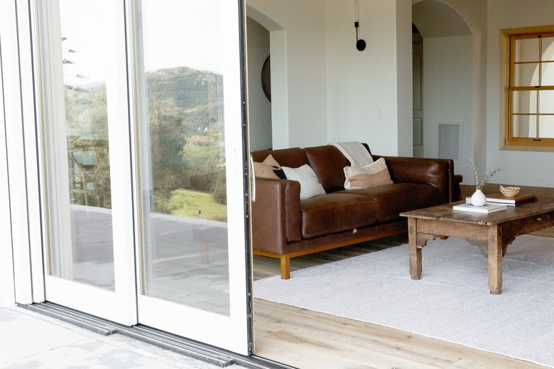 Glass sliding doors with painted-white frame. The doors are mostly open, leading into a living room. The walls of the living room are white. There is a window with a light wood frame on the opposing wall. In the living room, there is a brown leather rug with some throw pillows along with a stained wood coffee table with a few books and a small ceramic vase. There is a white rug on the floor.