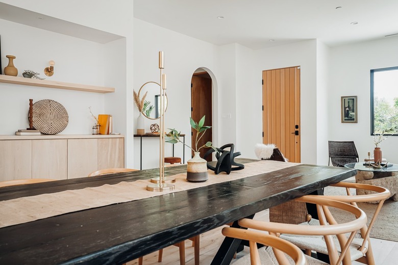 Black dining room table with a beige table runner, candlesticks, twisted globe sculpture, and beige wood chairs. A wood sideboard with pottery.