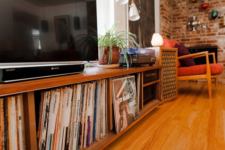 A wood credenza with a record collection, tv and a plant, in a living room with a brick wall and wood floor.