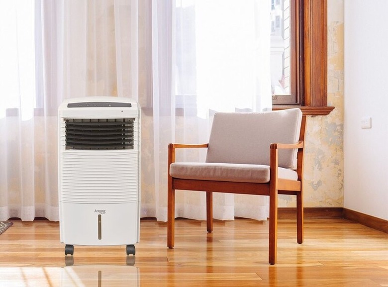 White portable air conditioner in a bright corner of a room with white sheer curtains, a beige upholstered and wood chair, and polished floors