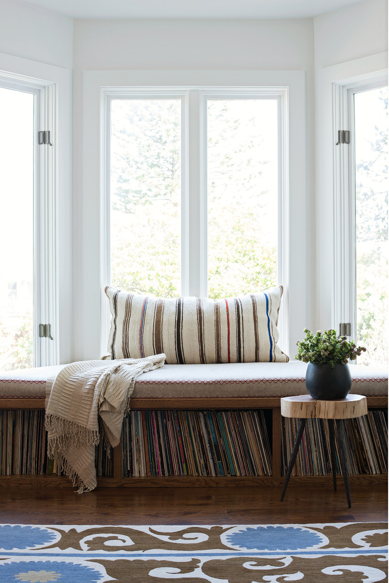 White bay window with pillow, a brown cushion, record player storage and wood side table with a plant.