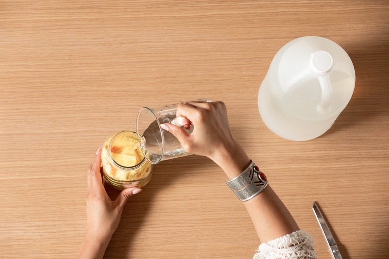 Hand pouring liquid soap into mason jar filled with wadded yellow dryer sheets next to plastic jug on wood countertop