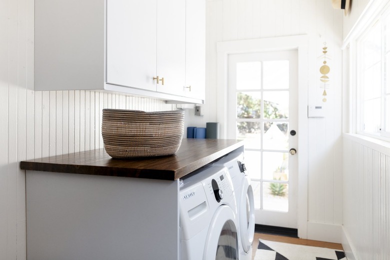 Wood tabletop with a woven basket, and a washer-dryer set, and white cabinets above.