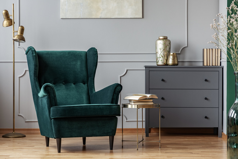 Books on small gold table next to green velvet wing back chair in grey living room.