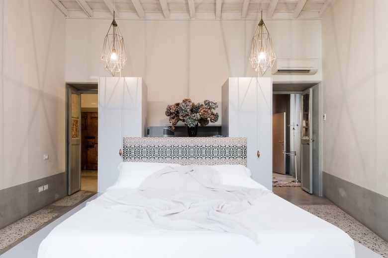 White bed against patterned headboard with potted plants with large hanging lights in white-walled room with doorways on either side