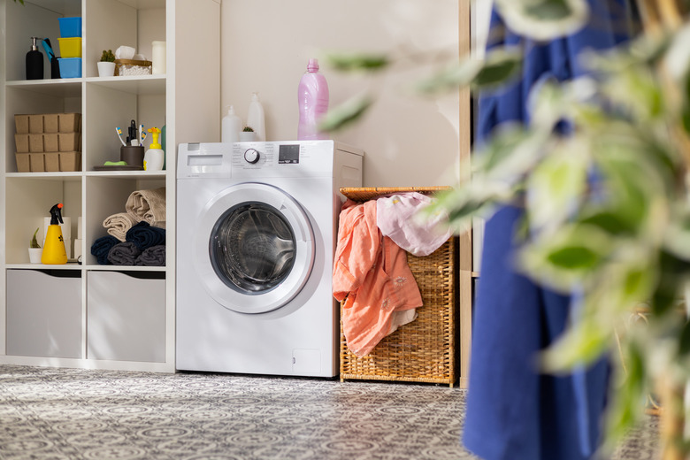 View of home laundry room, shelf with detergent, powder, towels, open washing machine with empty drum, next to wicker basket with colorful laundry items.