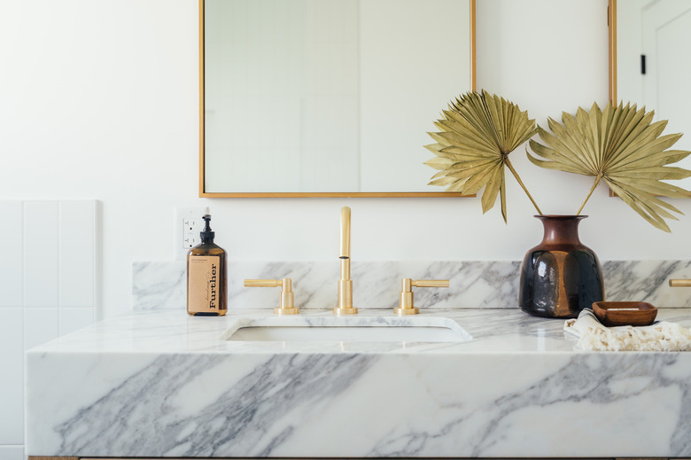 marble vanity countertop with ceramic undermount sink, gold faucet and handles, brown soap dispenser, vase with paper flowers, rectangular mirror with gold trim