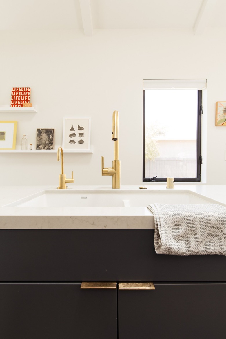 An undermount sink with gold faucet hardware. Black cabinets with tab handles. Framed artworks on wall shelves and a window.