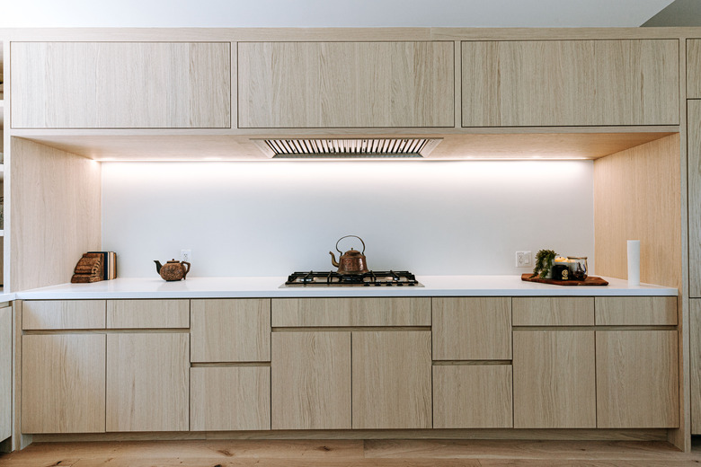 Minimalist kitchen with light wood cabinets, and a white countertop with a small stovetop and teapots of various styles