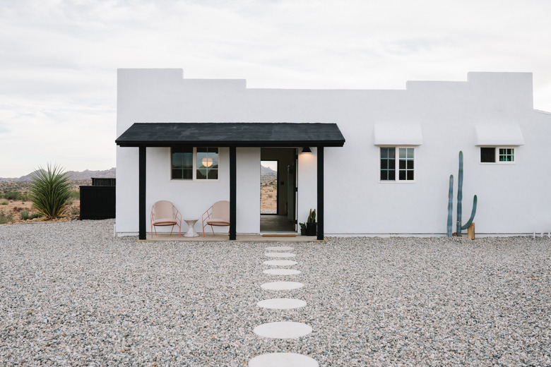 A stone path in the middle of a gravel walkway; a white southwestern-style home with a black awning