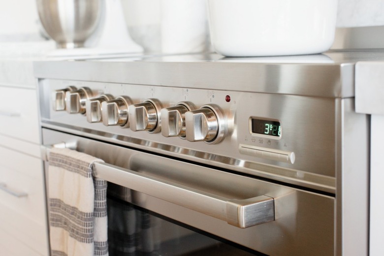 Closeup of an oven door and temperature control knobs on a stainless steel range. A white and grey striped kitchen towel is hanging over the handle over the oven door.