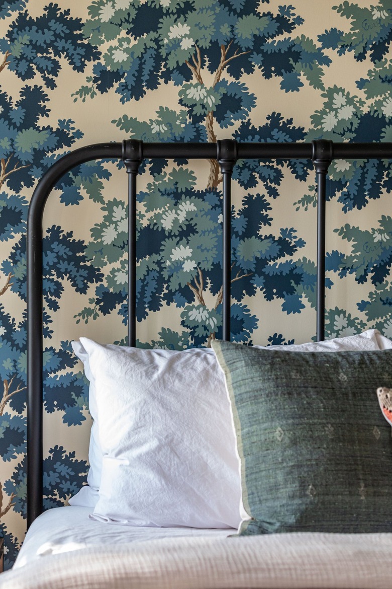 Bed with curved, black bar headboard, white bedding with a green throw pillow, and vintage blue tree wallpaper.