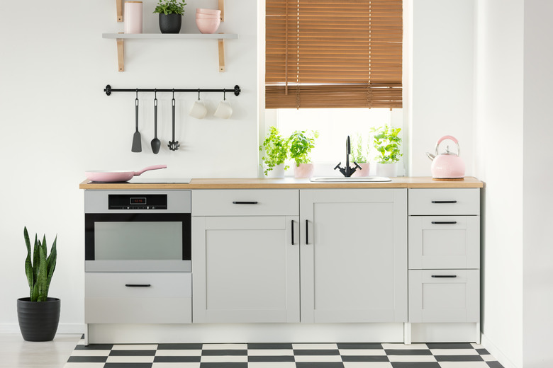 Real photo of a kitchen interior with checkered floor, white cupboards, pink accessories, plants, and window blind.