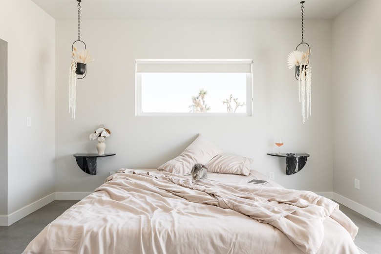 Minimalist bedroom with beige bedding, twin black shelf nightstands, double hanging planters, and rectangular window