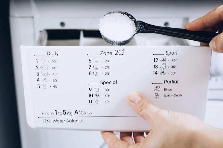 Hand using black measuring ladle to pour powder soap into second compartment of detergent compartment in white washing machine