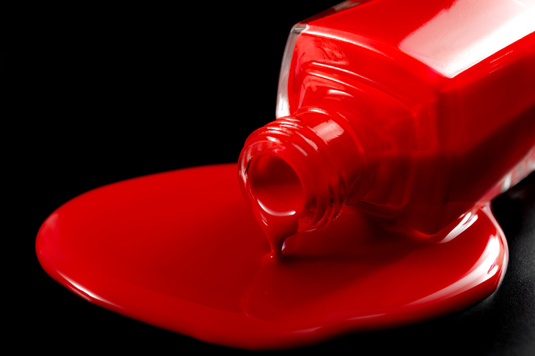 Bottle of red nail polish sitting in a pool of spilled nail polish on black background with dramatic lighting.