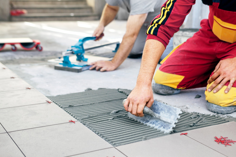 Ceramic Tiles. Tiler placing ceramic wall tile in position over adhesive, with handy man in background cutting ceramic tile.