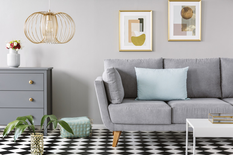 Light blue pillow placed on grey couch in bright living room interior with checkerboard linoleum floor, fresh flowers in vase on cupboard two posters hanging on the wall and gold lamp