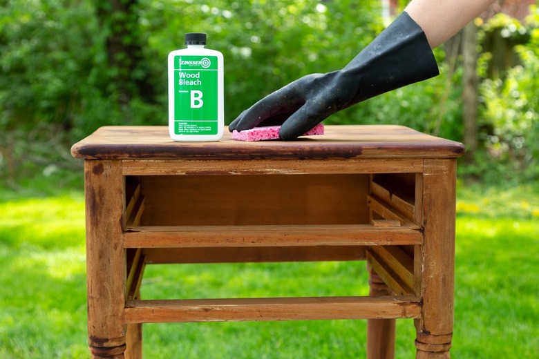 Wood side table without double drawers, and a gloved hand applying bottled wood stain to the tabletop with a sponge