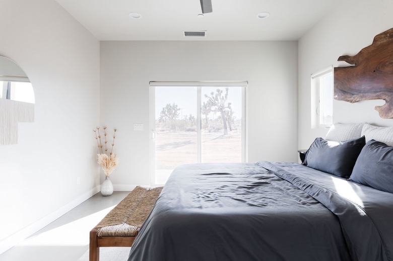 Minimalist desert bedroom with gray bedding, wood bench, organic wood art, half-dome fringe mirror, dried florals