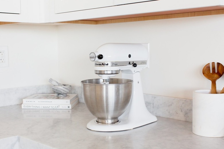 White cabinets above a marble counter. On the counter there is a white KitchenAid stand mixer with a stainless steel mixing bowl. Next to the mixer is a white utensil holder with wooden salad tongs. In the corner are two stacked cookbooks – 