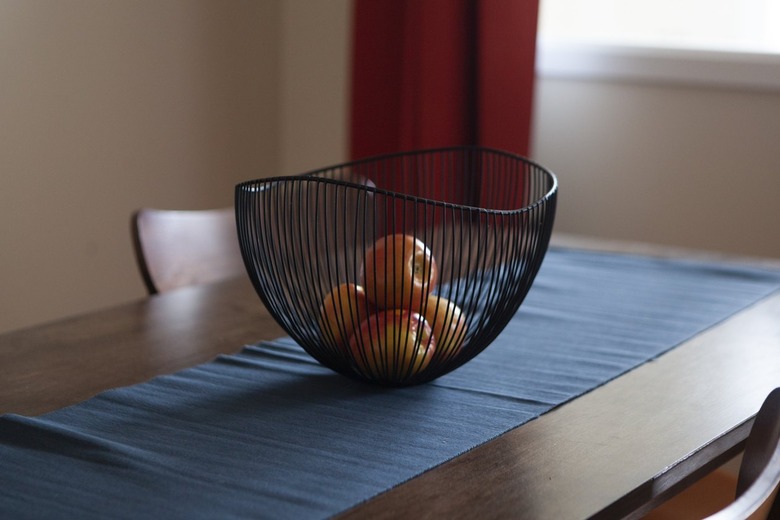 A wood table with a blue table runner, and a black wire bowl with apples.