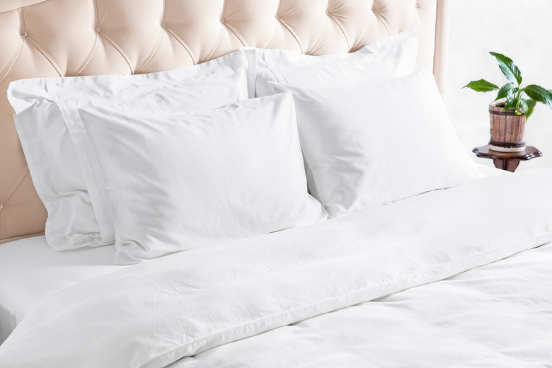 Bedroom with bed, white bedding, and bedside table. White pillows, duvet and duvet case on bed with upholstered beige headboard.