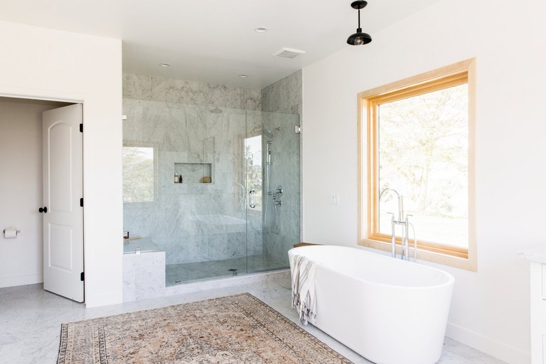 Bathroom with a freestanding bathtub, marble/granite shower with glass doors,  bell pendant light, neutral rug, and wood frame window.