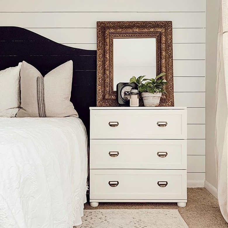 Off-white three-drawer dresser with a gold mirror and plant on top, next to a bed with a black headboard and white bedding