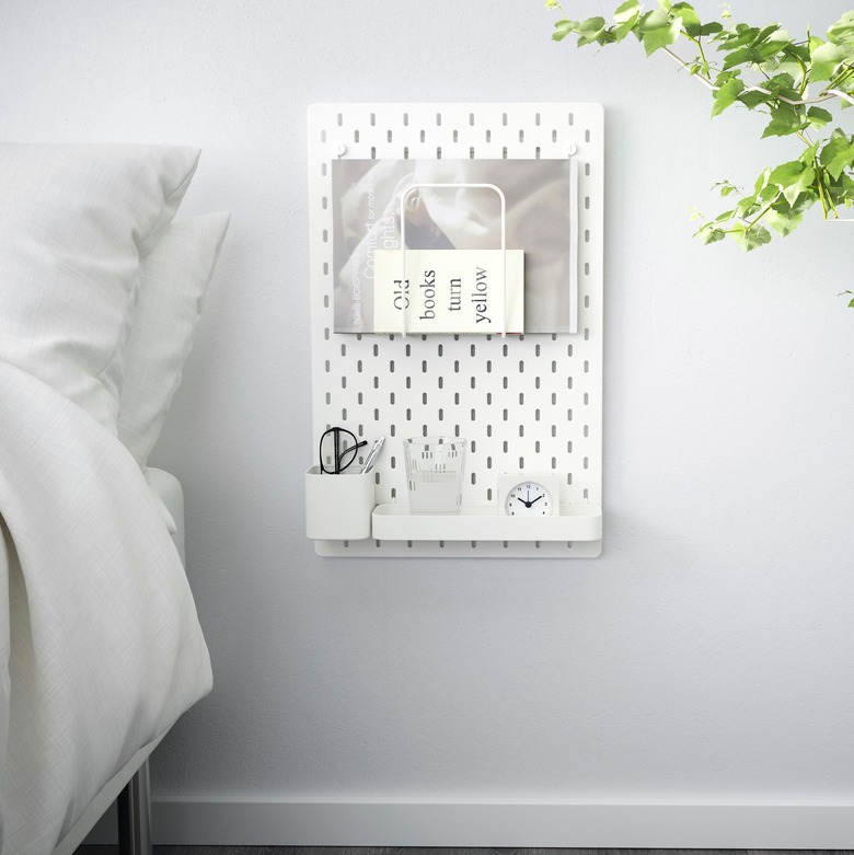 IKEA bedroom idea with a white pegboard mounted next to a bed. Pegboard has modules for glasses, water, an alarm clock, and books