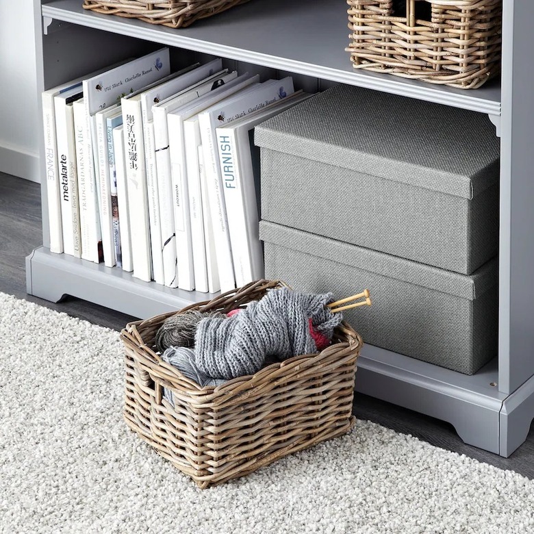 A woven basket full of knitting supplies on a gray carpet in front of a gray bookshelf.