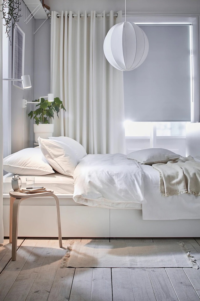 A white bed with white bedding in front of white curtains surrounded by decor like hanging lights, plants, and a wooden side table.