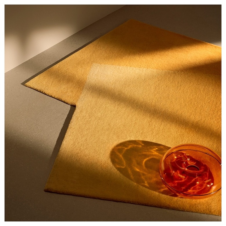 An orange VARMBLIXT Ikea serving bowl rests on the floor next to two small orange rugs overlapping each other. The bowl looks like a donut, and the light that's reflecting off of the bowl creates a beautiful light-filled shadow.