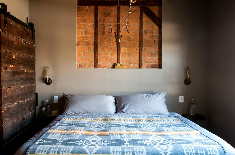 industrial farmhouse bed with a blue bedspread in a room with one wall sided with wooden planks and one in drywall with a cutout exposing brick