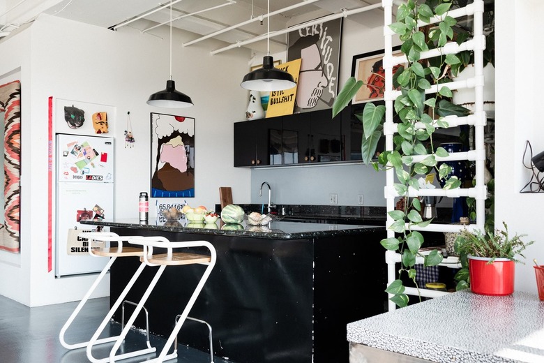 An industrial kitchen with black cabinets, black pendant lights, curved wood stools, and a pvc pipe shelf. Decorated with art and plants.