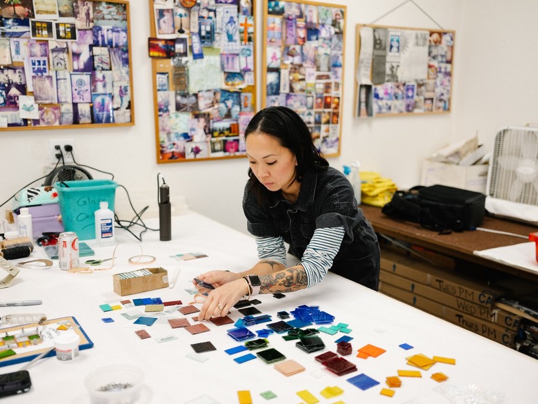 Asian woman with medium-length black hair with tattooed arms wearing black top with blue and white striped sleeves in art studio over workspace with colorful stained glass