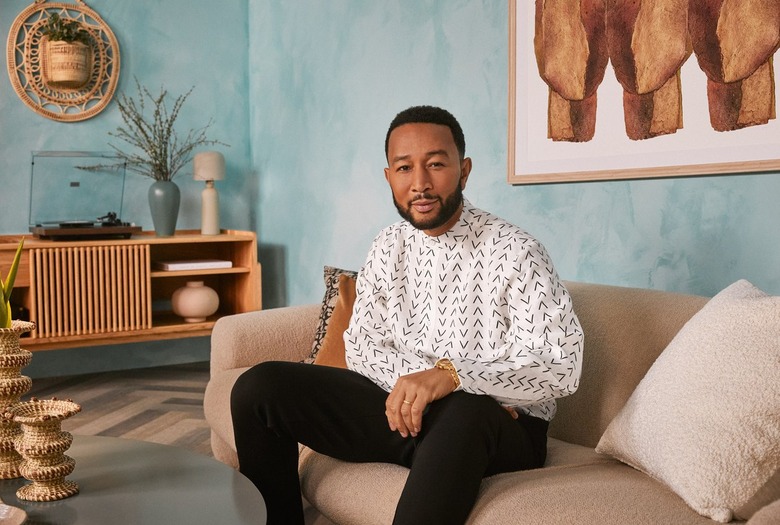 Musician John Legend sits on a couch wearing black pants and a white and black button-up shirt.