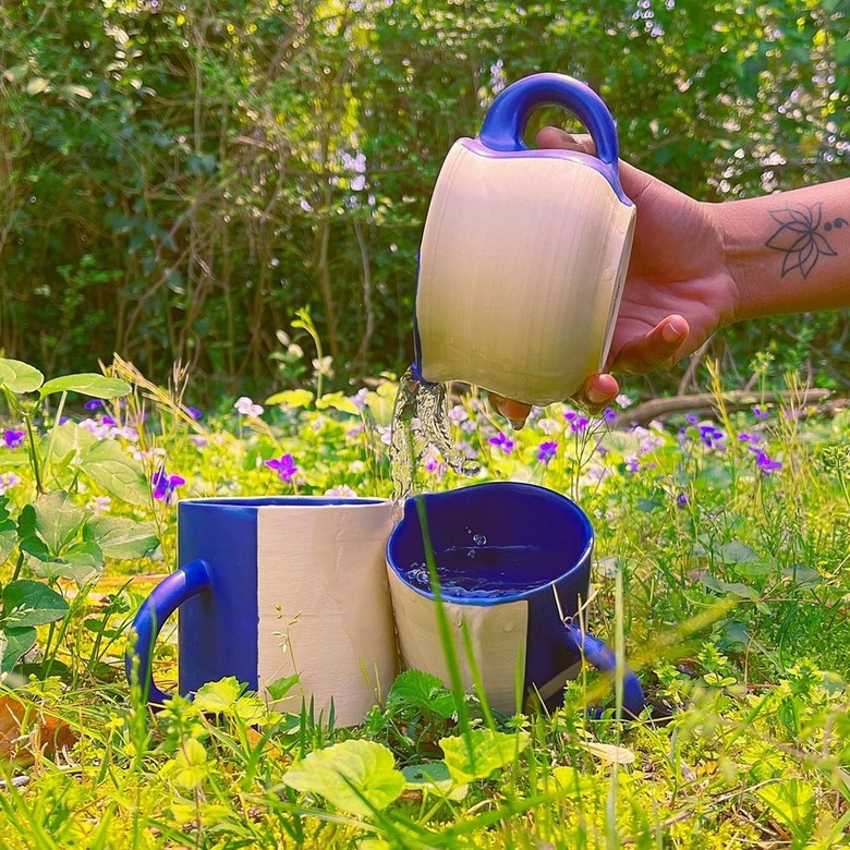 two ceramic mugs in the grass, another mug held by someone's hand on the side