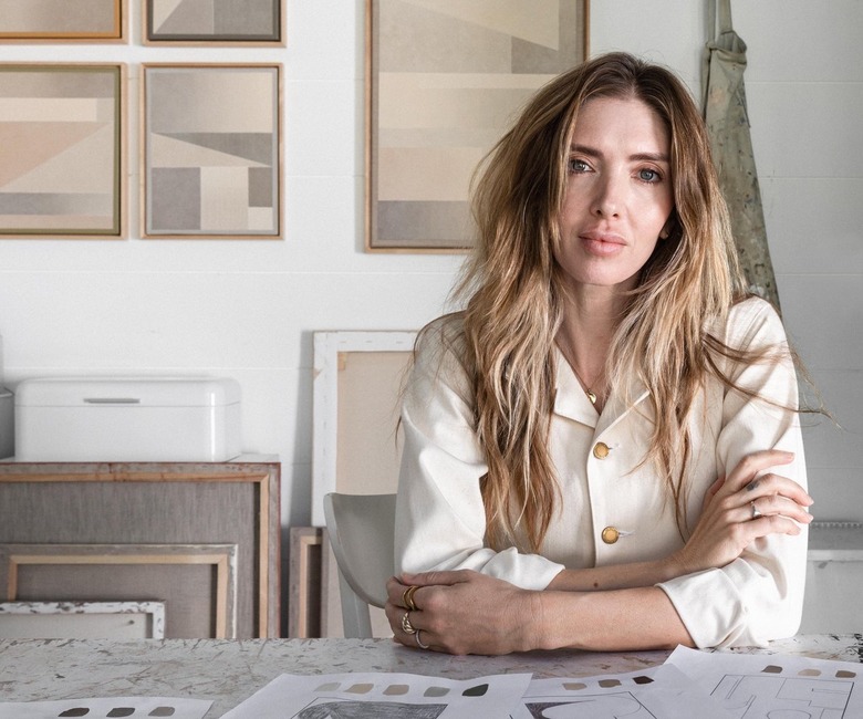 Karli Henneman in a white shirt with buttons leaning on a table with her paintings in the background on a white wall.