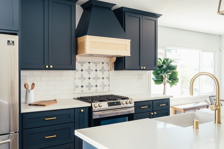 Dark blue kitchen cabinets, ornate backsplash by stovetop, white kitchen island countertop with gold sink faucet.